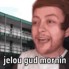 a man with a beard is making a funny face in front of a building with the words `` jelou gud mornin '' .