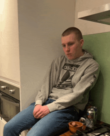 a man wearing a grey adidas sweatshirt sits on a kitchen counter