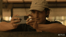 a man wearing a hat is taking a picture of himself with his phone with a netflix logo in the corner