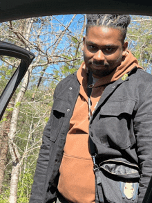 a man wearing a black jacket and a brown hoodie stands in front of a car