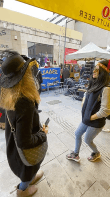 two women standing on a sidewalk in front of a sign that says ' tadex ' on it