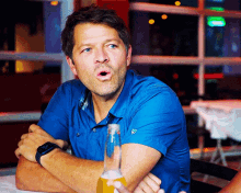 a man in a blue shirt is sitting at a table with a bottle of beer in front of him