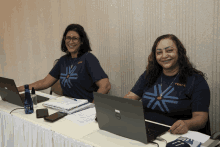 two women are sitting at a desk with laptops and one is wearing a shirt that says pen / a