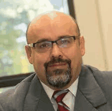 a bald man with a beard wearing glasses and a suit and tie is looking at the camera .