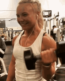 a woman in a white tank top is lifting a dumbbell in a gym with a reebok machine in the background