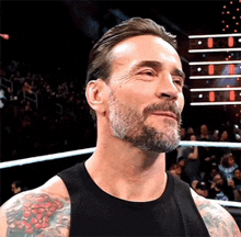 a man with a beard and tattoos is standing in a wrestling ring in front of a crowd .