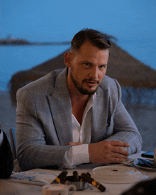 a man in a suit sits at a table with cigars in front of him