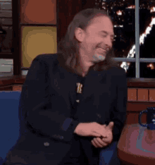 a man with long hair and a beard is smiling while sitting at a table with a mug on it .