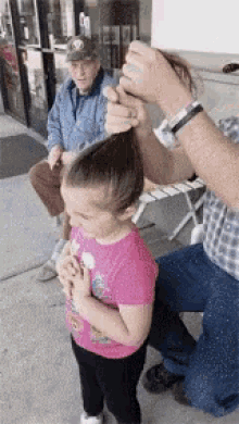 a little girl in a pink shirt is getting her hair braided by a man in a plaid shirt .