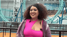 a woman with curly hair wearing a pink top and a purple jacket smiles in front of a roller coaster