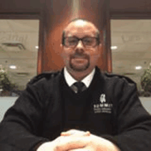 a man with a beard and glasses is sitting at a table with his hands folded in front of a window .