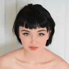 a woman with short black hair and bangs is standing in front of a white wall and looking at the camera .