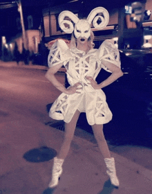 a woman wearing a ram mask and a white dress stands on the sidewalk