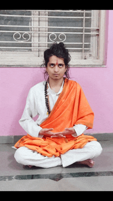 a man in a white shirt and orange robe sits in front of a pink wall