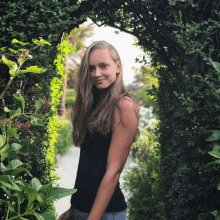 a woman in a black tank top stands in a lush green garden