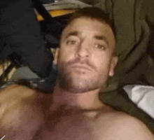 a shirtless man with a beard is laying on a bed taking a selfie .