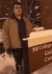 a man in a jacket is standing in the snow next to a sign that says `` 300 college guild college '' .