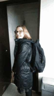 a girl wearing glasses and a black backpack is standing in a doorway