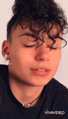 a young man with curly hair is wearing a black shirt and a necklace and looking at the camera .