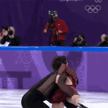 a man in a red jacket is taking a picture of a figure skater in front of the olympic rings