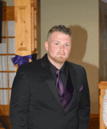 a man in a black suit and purple tie stands in front of a wooden pillar