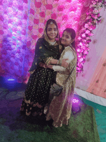 two women pose for a picture in front of a wall of flowers