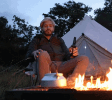 a man sits in front of a tent holding a bottle of beer that says ' beer ' on it