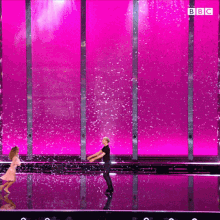 a man and a woman are dancing on a stage in front of a pink wall that says bbc on it