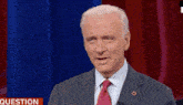 a man in a suit and tie is standing in front of a red curtain and a sign that says question