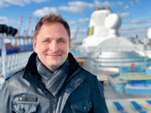 a man wearing a scarf and a leather jacket smiles in front of a large ship