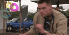 a man in a tan jacket and tie is sitting in front of a blue car and inflatable donuts .