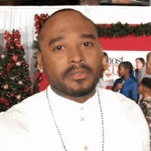 a man with a beard is wearing a white shirt and a necklace while standing in front of a christmas tree .