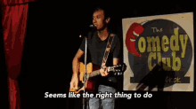 a man singing into a microphone while playing a guitar in front of a sign that says the comedy club