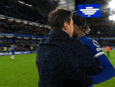 two men hugging on a soccer field with a blue sign in the background that says ' everton ' on it