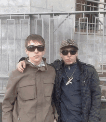 two young men are posing for a picture in front of a fence . one of the men is wearing sunglasses and a hat .