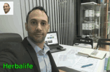 a man in a suit sits at a desk in front of a laptop with the word herbalife on the bottom right