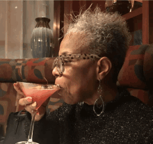 a woman wearing glasses and earrings drinks a martini