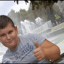 the boy is giving a thumbs up in front of a fountain .