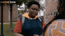 a woman with glasses and a backpack talks to another woman in front of a building with the showtime logo behind her