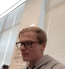 a young man wearing glasses is sitting in front of a window with blinds .