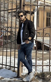 a man leaning against a fence wearing sunglasses and a black jacket