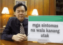 a man is sitting at a desk holding a sign that says `` mga sintomas na wala kang utak ''