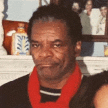 a man wearing a red scarf and a black shirt is standing in front of a fireplace .