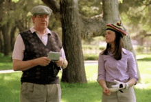 a man and a woman are standing next to each other in a park . the woman is wearing a hat .