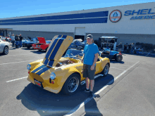 a man stands in front of a yellow car with the hood open in front of a building that says shel american