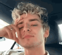 a young man with curly hair is sitting in the back seat of a car with his hand on his forehead .