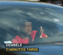 a man in a red shirt is driving a car with the words dembele 11 minutos tarde on the bottom .