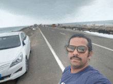 a man wearing sunglasses stands in front of a white car on the side of a road