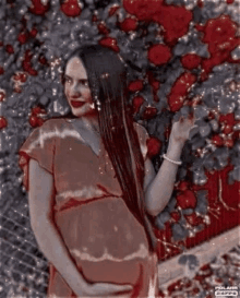 a pregnant woman in a red dress is standing in front of a bush of red flowers .