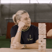 a woman wearing a tezis shirt sits at a table with a stack of wooden blocks